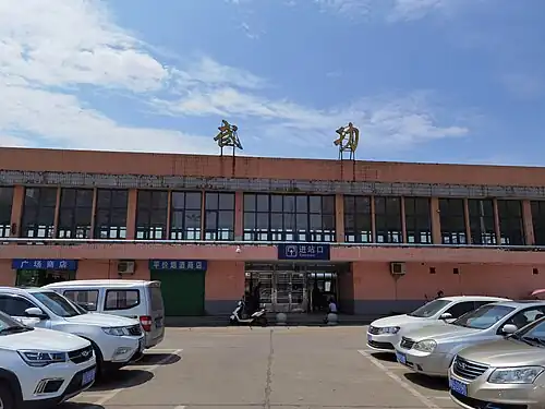 A two story high concrete building. The first floor lacks windows, while the second floor has floor-to-ceiling windows. In the center, on the first floor, there is an entrance. In the foreground, there is a parking lot.
