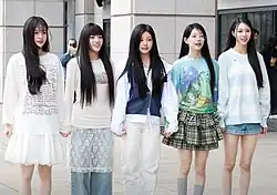 Five women standing in a row outdoors, holding hands and smiling. They are dressed in casual, stylish outfits featuring soft and pastel colors.