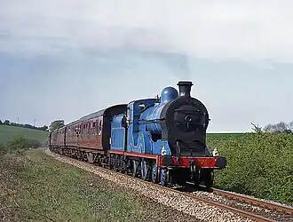 Great Northern Railway number 171 pulls a train of passenger coaches along a track through a field. The locomotive and tender are painted blue with gold lines, and red trim on her running boards; her smokebox and buffers are painted black. The coaches behind her are painted a reddish-brown, with black roofs.