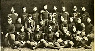 A sepia-tone picture of around thirty young white men, all in American football uniforms from the beginning of the 1900s. They are taking a team picture, staged in rows and all facing directly into the camera. Some are sitting cross-legged.