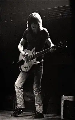 This photo is in black and white. Malcolm, aged 29, is shown in left profile, playing guitar with both hands. He has over shoulder-length hair, and is wearing a dark T-shirt, jeans and shoes.