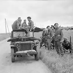A black and white photograph of a jeep with soldiers sitting on top and standing beside it. The soldiers sitting on the jeep are three German soldiers and one British soldier who is interrogating the Germans. On the bonnet of the jeep is small motorcycle, while in the background is a Horsa glider