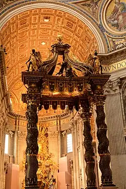 Photo shows the baldachin standing in the centre of the church, viewed looking towards the nave. There is an altar beneath it which has a red and gold frontal cloth decorated with large crosses.