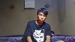 Didit staring at the camera with a dull expression wearing a Justin Bieber T-shirt, at his dirty bedroom with one purple bed.