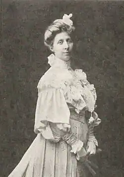 A white woman, standing, hands clasped around a fan held low in front, wearing a ruffly white gown with a high neckline and large sleeves, with hair in a bouffant updo