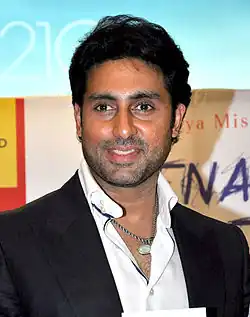 Abhishek Bachchan smiling in a black suit with an open-collared white shirt and layered necklaces at a public event.