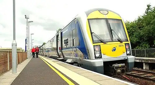 An image of a Northern Ireland Railways train pulled into the station with a view down the platform with two people getting off the train in the distance.