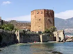 A eight-sided medieval tower built of red and yellow brick rises above a green sea in which swimmers play. Stone walls run along the shore and further up from the tower.