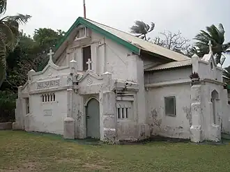 A picture of a small white church with spires, nestled next to palm trees and bushes.