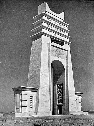 Black and white photograph of a large modernist stone arch in three-quarters view, set in a desert landscape. The bright white arch is pyramidal in shape, reminiscent of Ancient Egyptian pylons, with a wide opening that stops approximately halfway up the arch. Within the opening, on the inside of the arch, a carved bas-relief can be seen. Above the opening is a three-tiered attic, inscribed with the Latin quote "Alme Sol possis nihil Urbe Roma visere maius" in block capitals, split over the three tiers. Below this quote is a bronze sculpture of a man set within a recess, representing one of the Philaeni brothers and appearing to writhe in pain. Two small square constructions jut from the either side of the arch at its base.