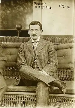 Black and white image of a man with a mustache, wearing a formal, Edwardian era suit and sitting with his legs crossed.