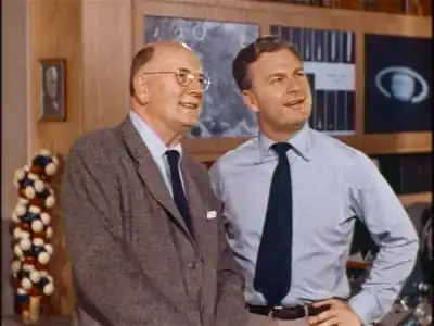 Photograph of two men standing. The man on the left is wearing a suit and tie; he is middle-aged, wears eyeglasses, and is bald. The man on the right is wearing a loose tie and is much younger. Behind the two men is a wood-paneled wall with photographs, one of which is the planet Saturn. There is also a large model of a molecule behind them, possibly illustrating DNA.