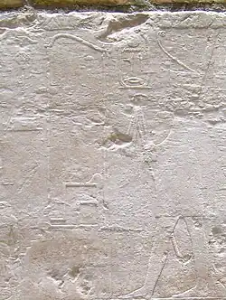 Relief showing a king seated on a throne, wearing a tight fitting dress and holding a flail. He is surrounded by hieroglyphs.