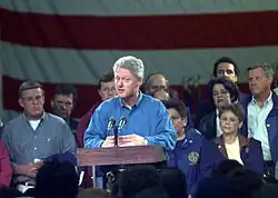 Bill Clinton is standing at a podium speaking to a crowd. The former mayor of Grand Forks is at the right of the image.