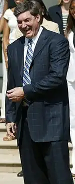 A man, wearing a black suit, is standing and laughing in front of spectators.