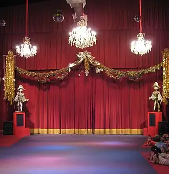A stage flanked by large marionette puppets with large chandeliers hanging above