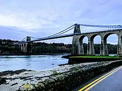 Pale blue metal suspension bridge carrying road traffic across a body of water with large stone arches at one end