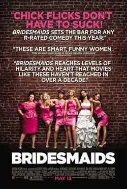 Against a red brick wall stands a sextet of women, five of them are wearing bridesmaid dresses, while the remaining one is wearing a wedding dress