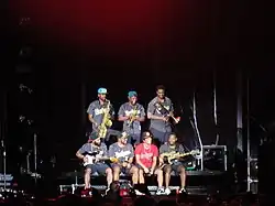A group of seven men sit and stand on a stage against a dark backdrop.