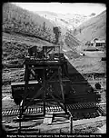 A photograph, taken outdoors, of men working on railroad tracks. There are two sets of tracks running horizontally and one set running up a hill to the distance. There is what appears to be a large chute over the two horizontal tracks. The vertical tracks are likely to be mining tracks with a coal car on them.