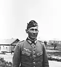A man wearing a side cap, military uniform with an Iron Cross displayed at the front of his uniform collar.