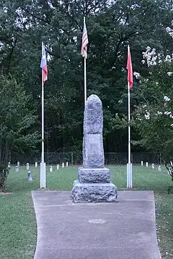 Photo of the 12-foot memorial stone obelisk erected in the center of the cemetery with three flagpoles in background containing flags of United States, Texas, and Arkansas