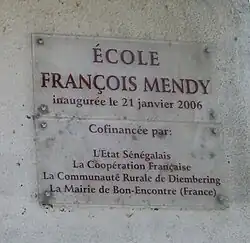 A clear plaque on a grey wall stating "École François Mendy" at the top with smaller writing below