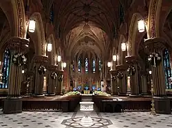 A church interior with a tall vaulted ceilings, wooden pews on a stone floor, and stained glass windows