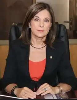 Caucasian female with long, dark brown, straight hair, wearing a dark jacket over a red top, sitting at a desk