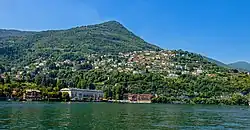 A mountain rising above water on a day with clear blue sky. There are large houses at the water's edge, and smaller houses higher up.