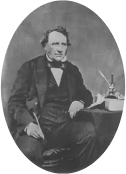 A middle aged Victorian gentleman sits beside a table wearing a dark suit with waistcoat and bow tie. His left elbow rests on the table. In his right hand he holds a quill pen.