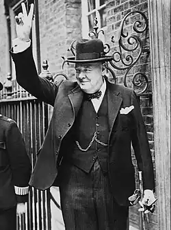 U.K. Prime Minister Winston Churchill in a black lounge suit with homburg hat and walking stick on Downing Street, giving his famous 'V' sign during World War II.