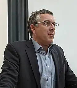 Bust of a clean-shaven white man with short gray hair, glasses, blue collared shirt, and dark gray suit jacket