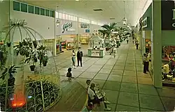 The interior of a shopping mall in the 1960s, with tiled floor. Visible are a fountain, several kiosk shops, planters with trees, and signage reading "Hanover Shoes", as well as several customers.