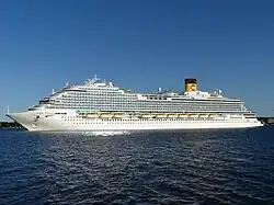 The port-side and aft of a cruise ship with white paint, many decks of balconies, and a yellow vertical cylindrical funnel with a blue 'C' painted on it, is shown from a distance.