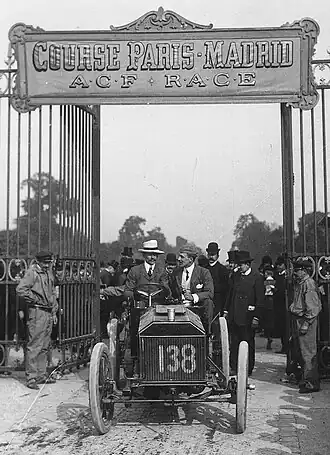 A poster of two men driving a car numbered 138 through an open gate. The poster is titled "Course Paris-Madrid - ACF Race"
