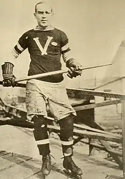 A man poses for a photo wearing skates and an ice hockey sweater, and holding a stick in his hands.