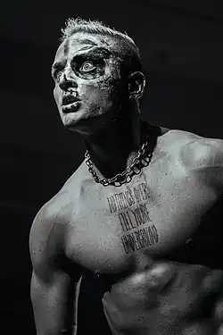A black and white photograph of a shirtless man with blonde hair, wearing face paint and a chain around his neck. A tattoo on his chest reads "Nothing's over til you're underground".