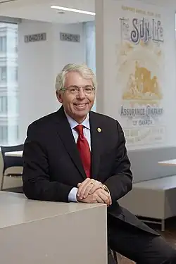 Colour photograph of Dean A. Connor, wearing a suit and red tie. he is sitting in front of a sign reading "the sun life assurance company"