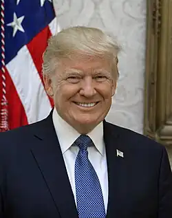 A head-and-shoulders portrait of Trump beaming in front of the U.S. flag, wearing a dark blue suit jacket with American flag lapel pin, white shirt, and light blue necktie.