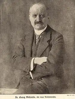 Black and white photo of an elderly man sitting in a suit with folded hands, mustache and bald head.