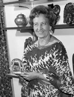 a woman dressed in African dress and jewelry holds a small sculpture of an African animal. Other artifacts line the shelves behind her.