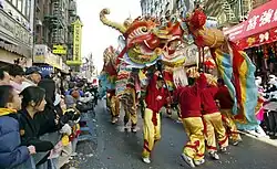 Several men in red and yellow outfits carry a colorful paper dragon in the street while onlookers watch behind police barriers.