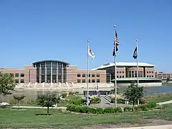Photo of two multi-story buildings which are connected. A parking garage can be seen in the background. A pond is in front of the principal building. A veteran's memorial, containing a sculpture, flagpoles, and well-manicured landscaping, is in the foreground.