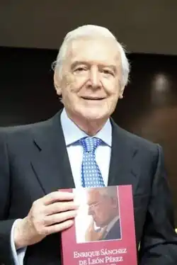 silver haired caucasian male with wearing a suit and sky-blue tie in a boardroom setting, showing off a book with red cover