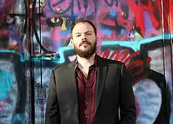 Erik Hoel wearing a burgundy shirt and dark blazer, standing in front of a wall covered in graffiti, looking sternly at camera with chin turned up and slight downward gaze