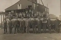 In a black and white photograph, group of 26 young to middle-aged men wearing suits and flat caps stand in two rows. Behind them is a large, industrial shed, and in the distance the roof of a house can be seen.