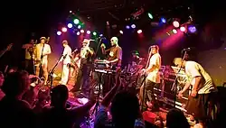 Seven musicians on a stage with various instruments and microphones, audience members raise their hands in a clapping motion