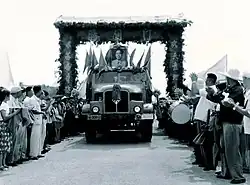 A truck decorated with an image of Mao Zedong passes through an arch, with crowds lining the road on either side