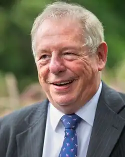 An older white man in a formal suit, with short, silver hair, smiling at someone or something out of shot.